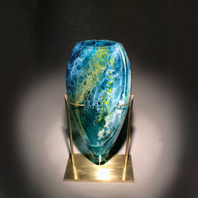 Sculpture d'un vase en verre soufflé travaillé à froid sur une base en laiton, de plusieurs teintes de bleu avec des touches de blanc et de jaune, réalisée par l'artiste Maryse Chartrand de Sutton, Québec, Canada.